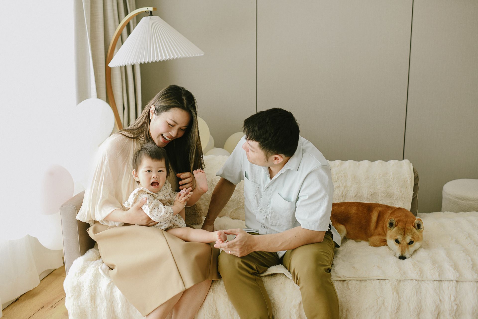 A family sits together on a couch with their baby and Shiba Inu dog in a cozy living room setting.
