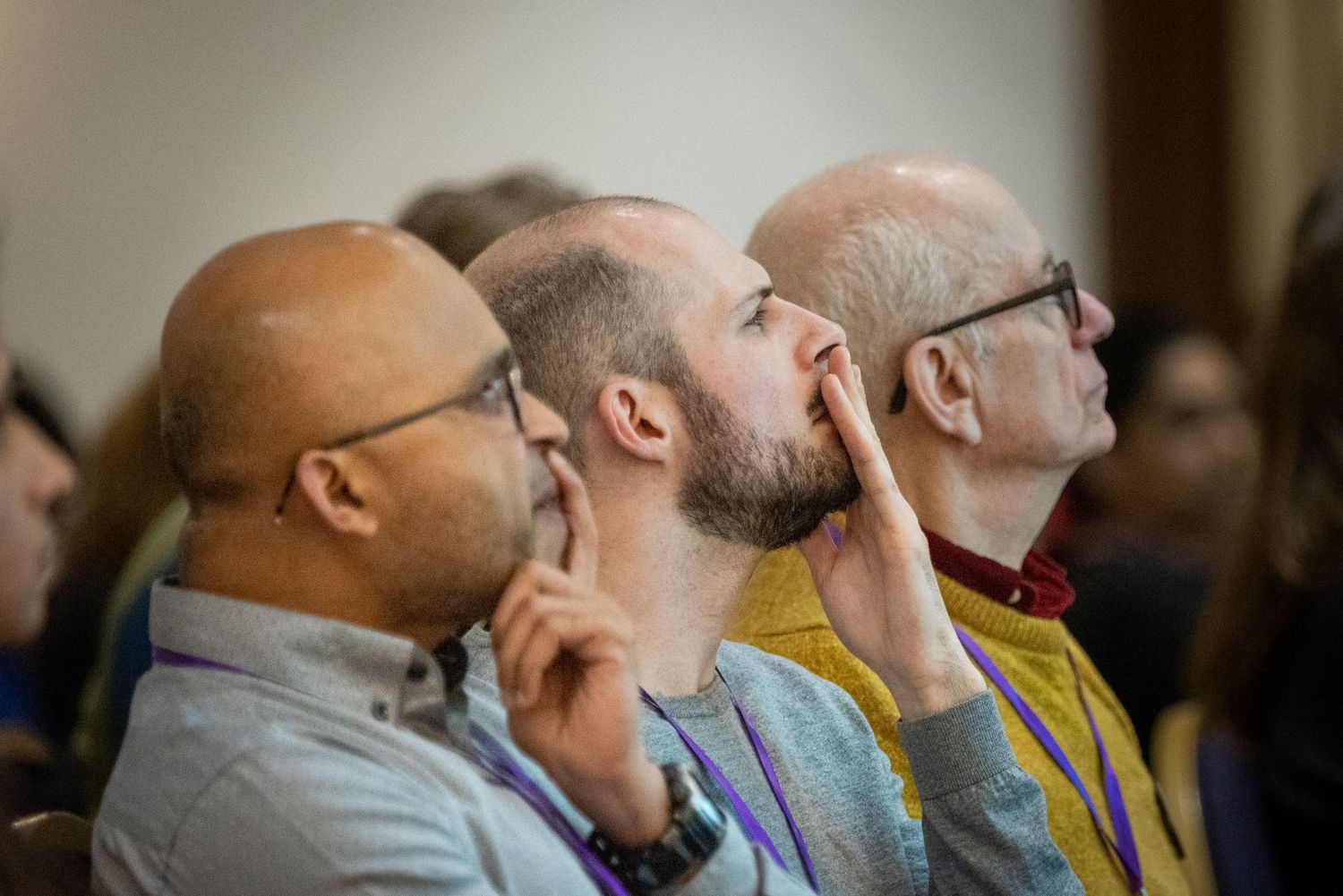 Three men sit attentively in an audience, one stroking his beard thoughtfully during a conference or seminar.