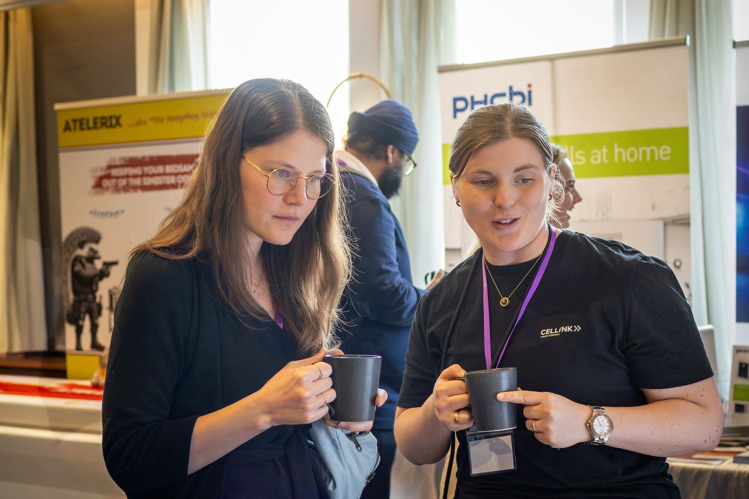 Two women holding mugs chat at an indoor conference event with vendor banners visible in the background.