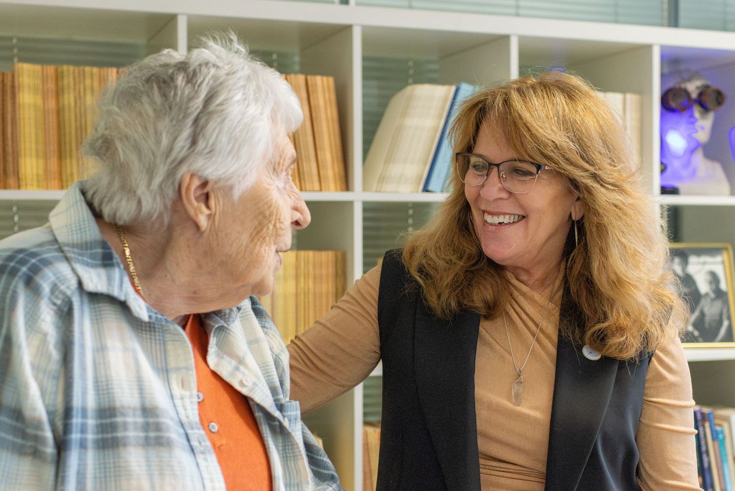 Two women smiling and conversing warmly in a library setting with bookshelves in the background.