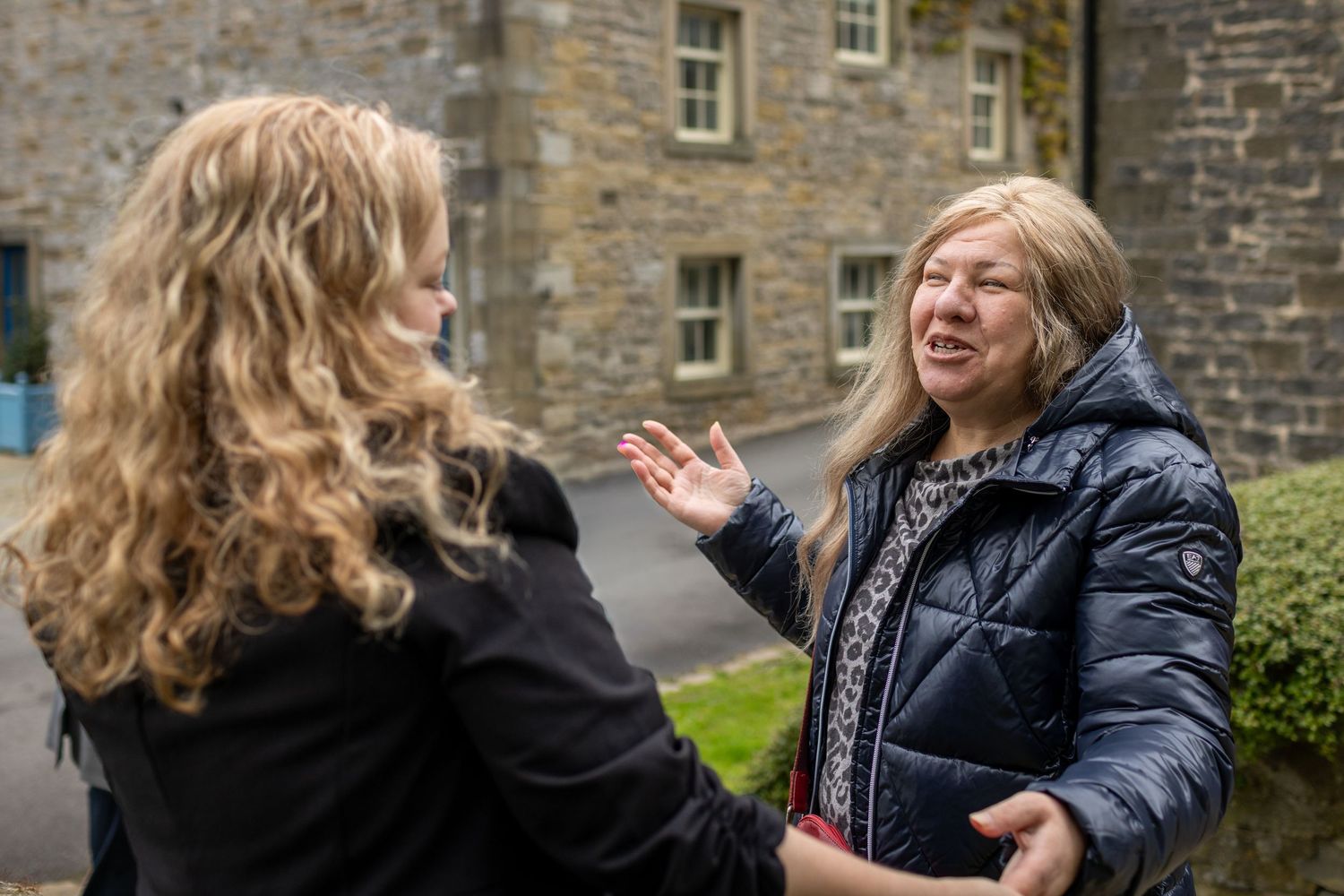 Two women having an animated conversation outdoors near a stone building.