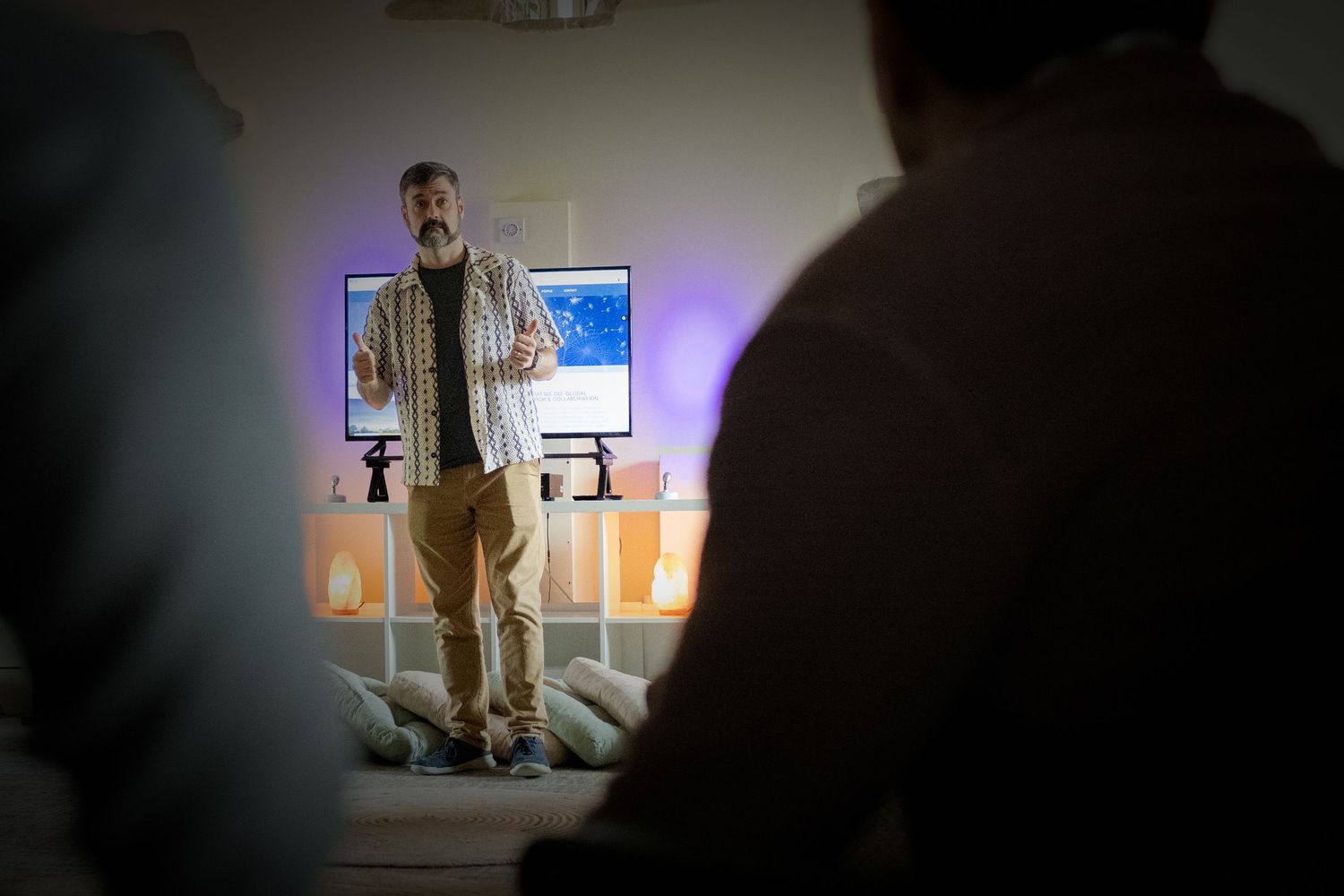 Man in plaid shirt presenting to audience in dimly lit room with colorful LED lights and monitor display.