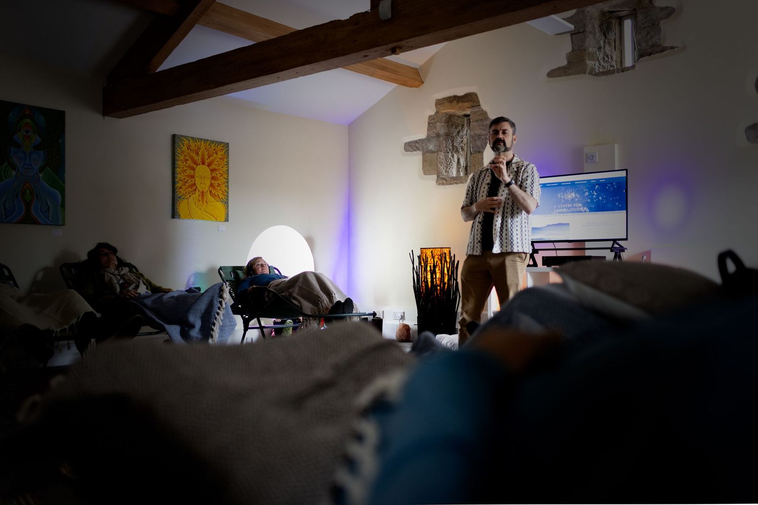 A man stands speaking in a dimly lit room while others relax on cushions during a sound healing session.