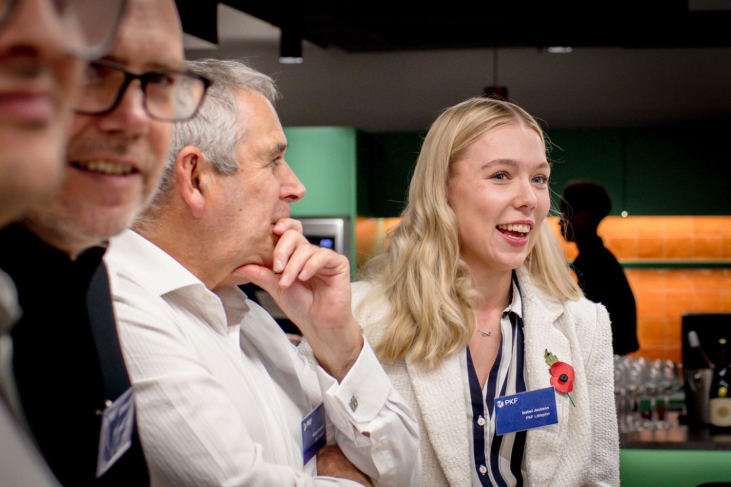 Professionals in white lab coats with name badges laugh together at a networking event with a green backdrop.