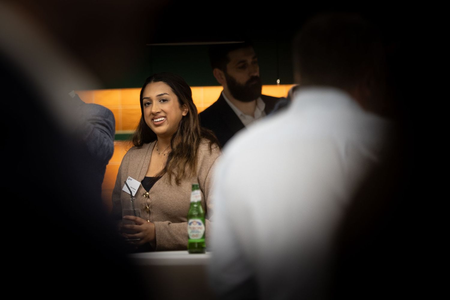 Woman smiling at social gathering with green Heineken bottle on table in dimly lit venue.