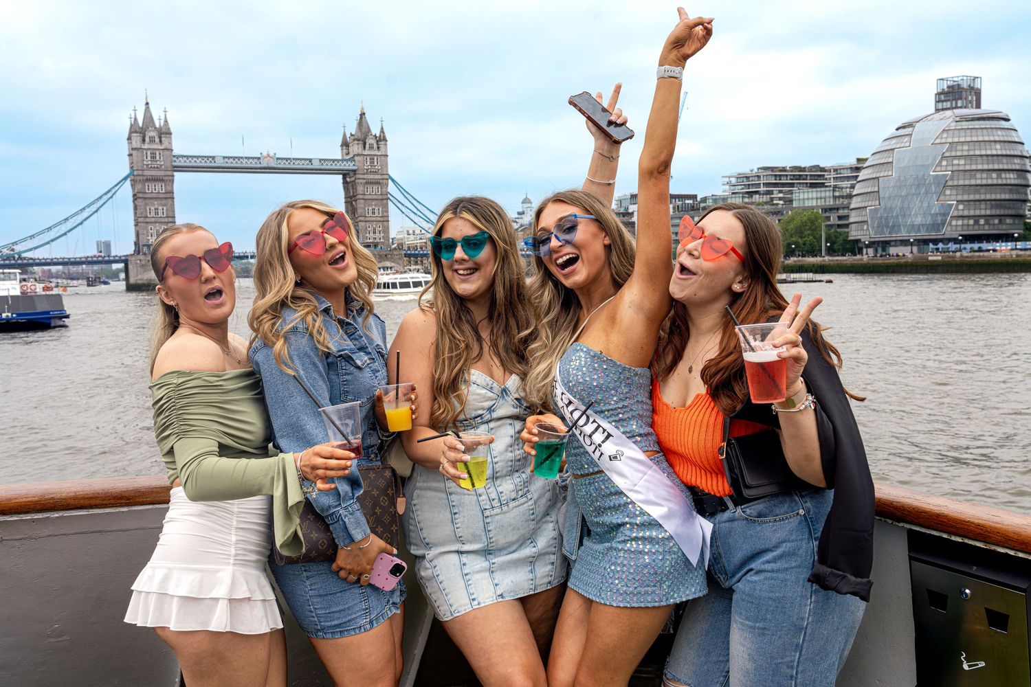 Group of friends enjoying drinks and laughing together on a boat tour along the River Thames with Tower Bridge visible.