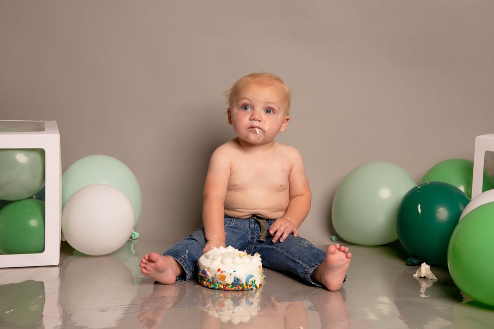 Baby sits on floor for cake smash photo shoot with green and white balloons and decorations in studio setting.