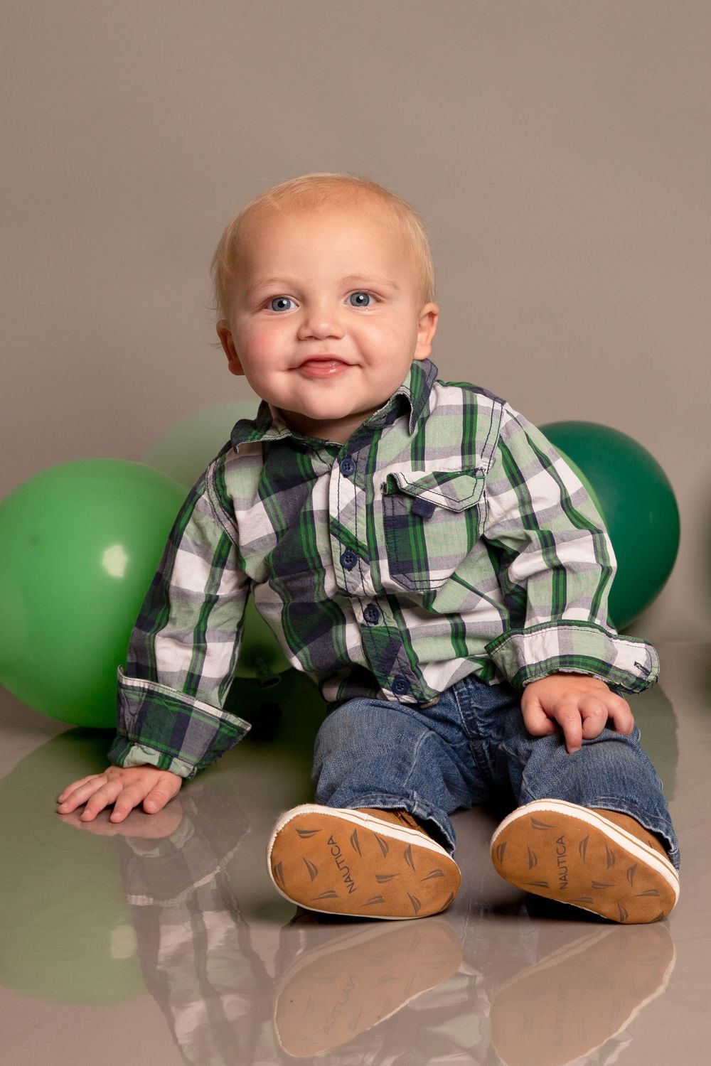 Baby in plaid shirt and jeans sits on floor with green balloons, wearing brown shoes and smiling brightly.