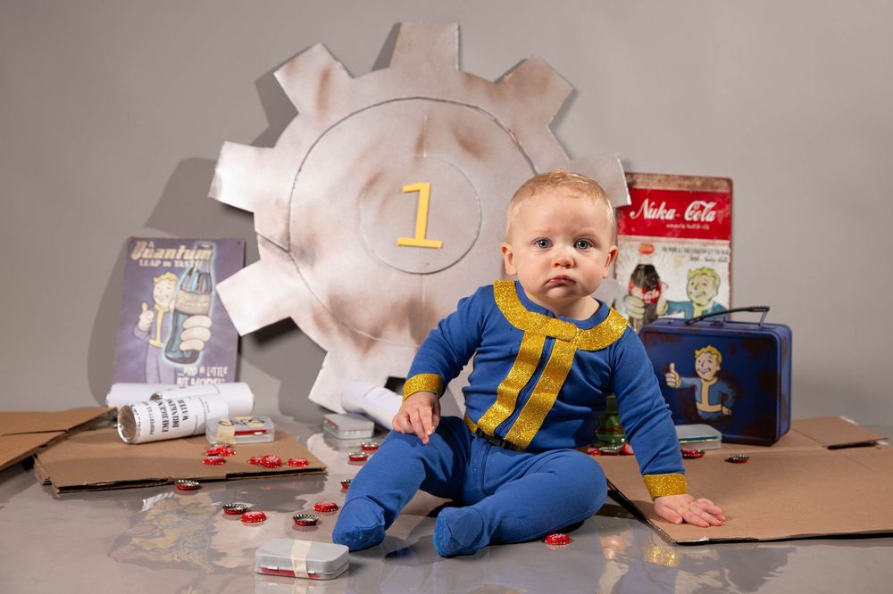 Baby in Fallout Vault Boy costume sits among Nuka-Cola props and cardboard for a themed first birthday photo.