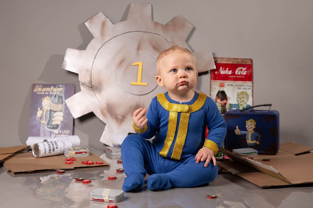 Baby in Fallout Vault Boy costume sits among Nuka-Cola props and cardboard for a themed first birthday photo.