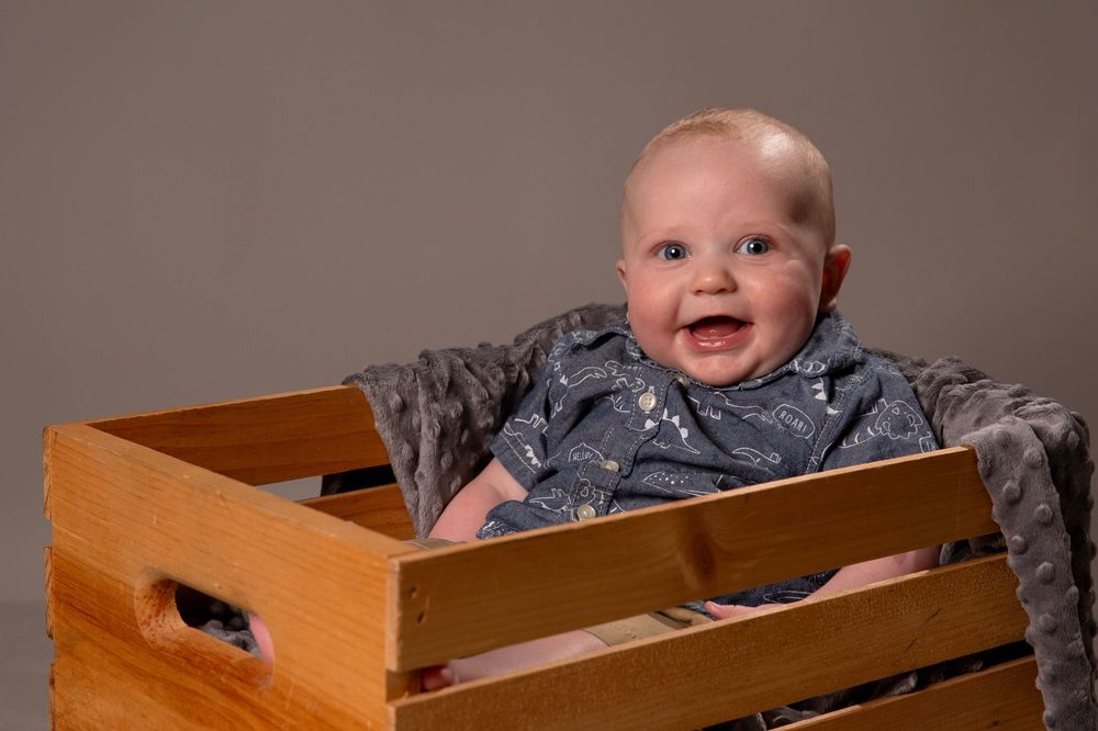A baby wearing grey clothing sits in a wooden box with a joyful expression.