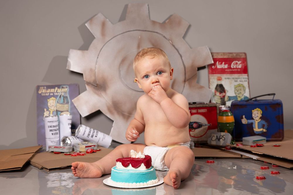 Baby in diaper sits with smash cake surrounded by Fallout game themed props including Nuka-Cola and vault boy items.