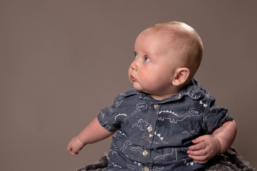 Baby in gray buttoned shirt gazes away from camera while sitting against neutral studio backdrop.