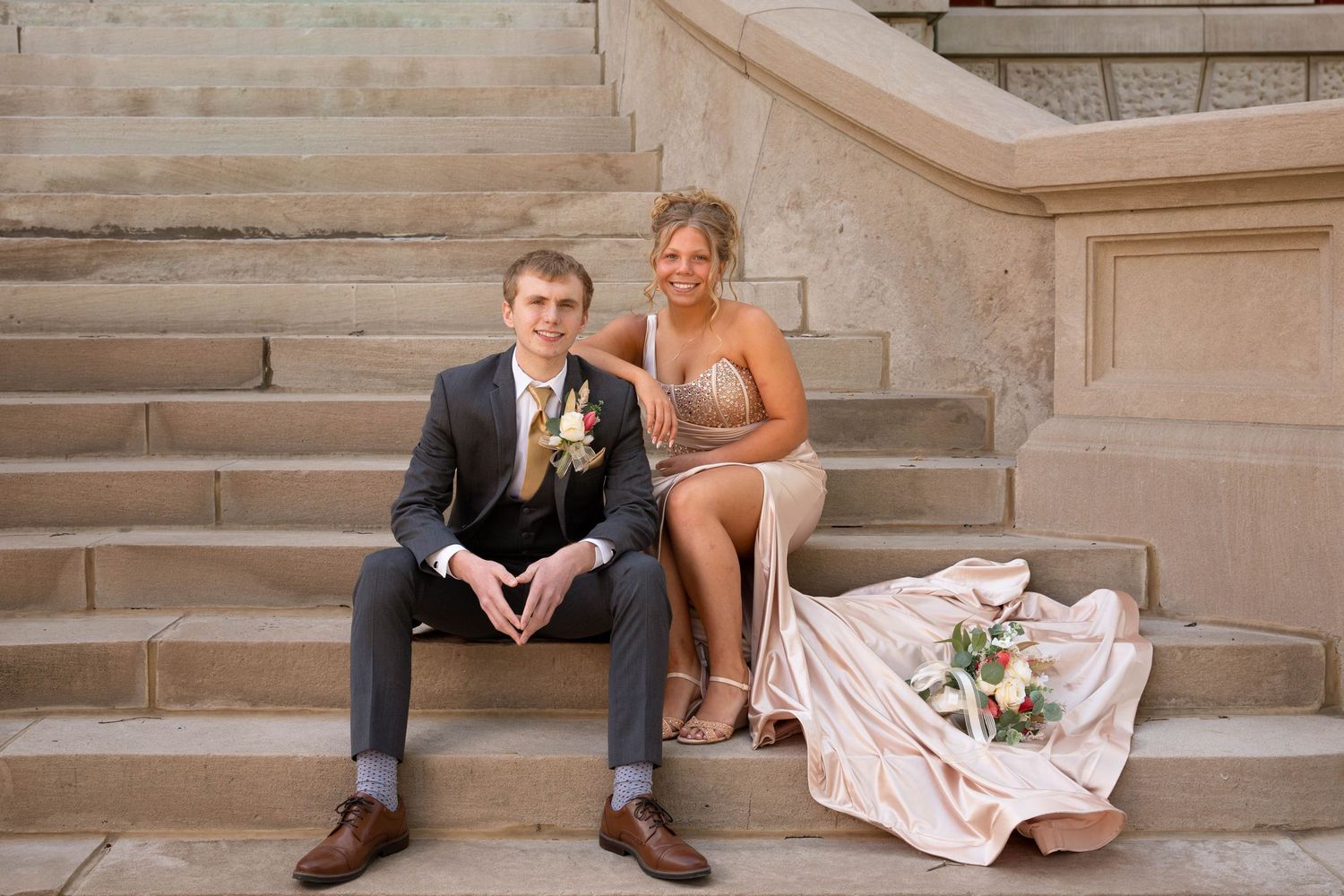 Two people in formal attire sit together on elegant stone steps with a bouquet of flowers beside them.