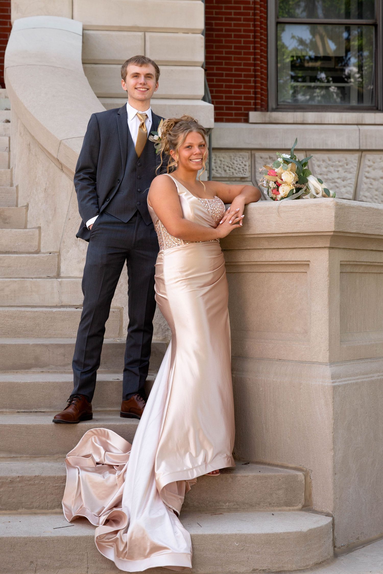 A couple in formal attire share a playful moment on stone steps outside a brick building.