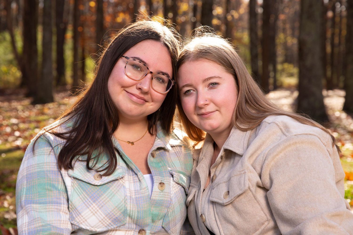 Two people smile warmly while sitting together outdoors in autumn as sunlight filters through trees in the background.