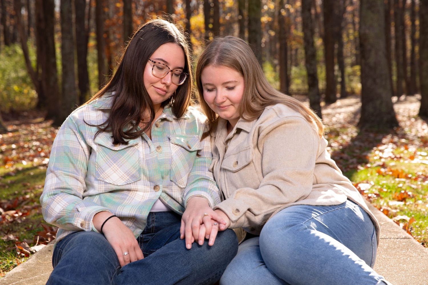 Two people share a heartfelt moment while sitting together on fallen autumn leaves in a sunlit forest setting.