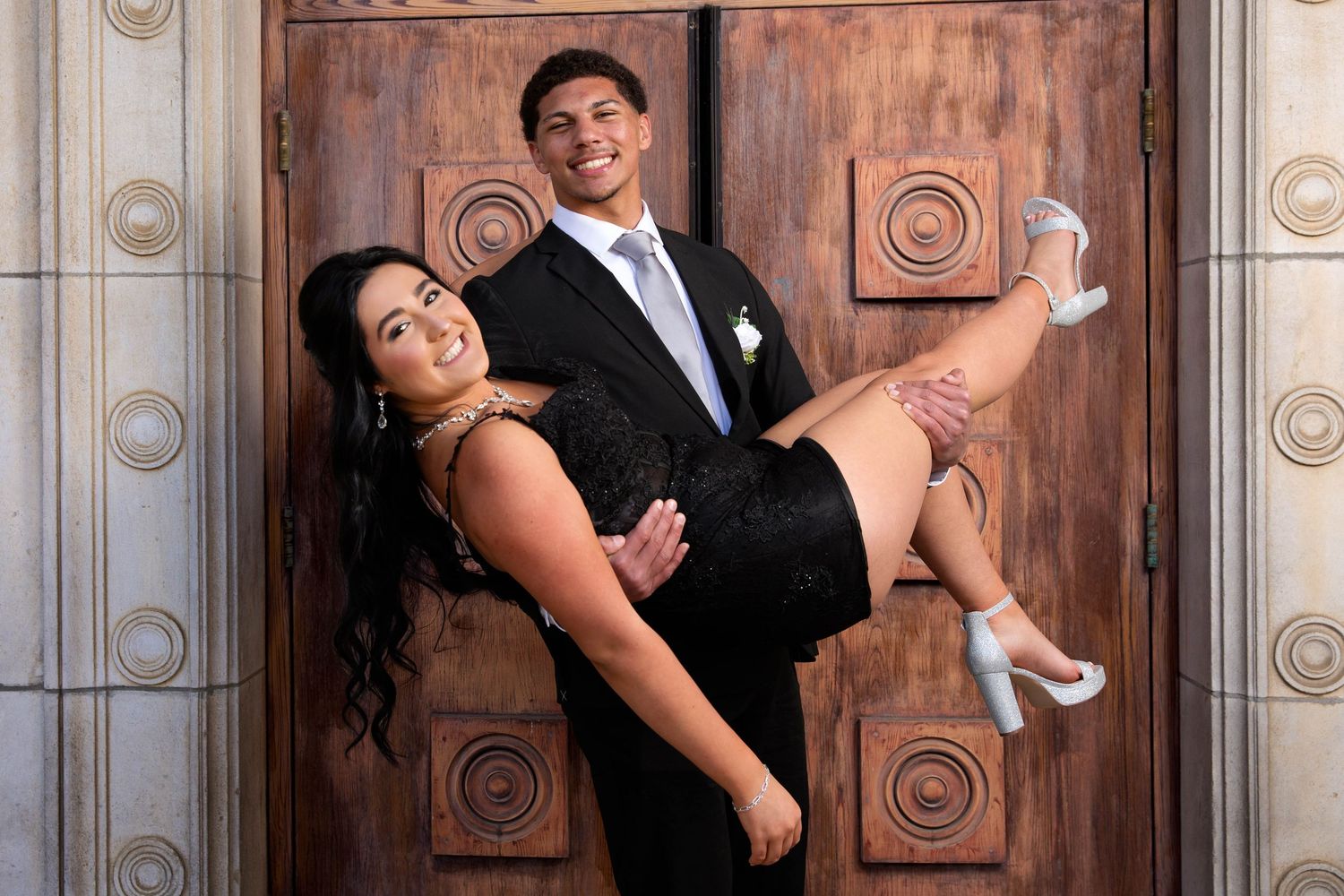 A formally dressed couple poses playfully against an ornate wooden door during what appears to be a special occasion.