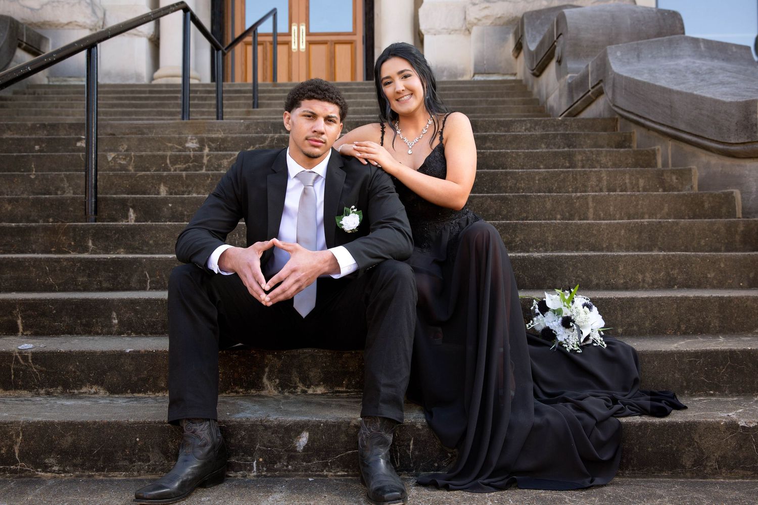A romantic couple sits on stone steps outdoors in formal attire with black and white colored clothing.