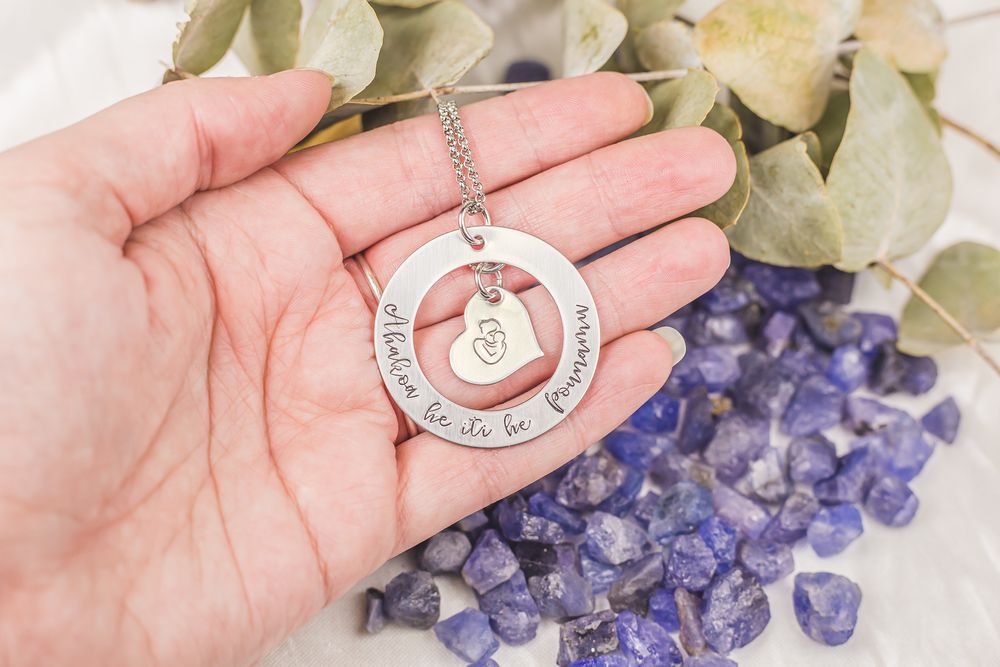 A silver circular pendant necklace displayed on an open palm with purple crystals and eucalyptus leaves in background.