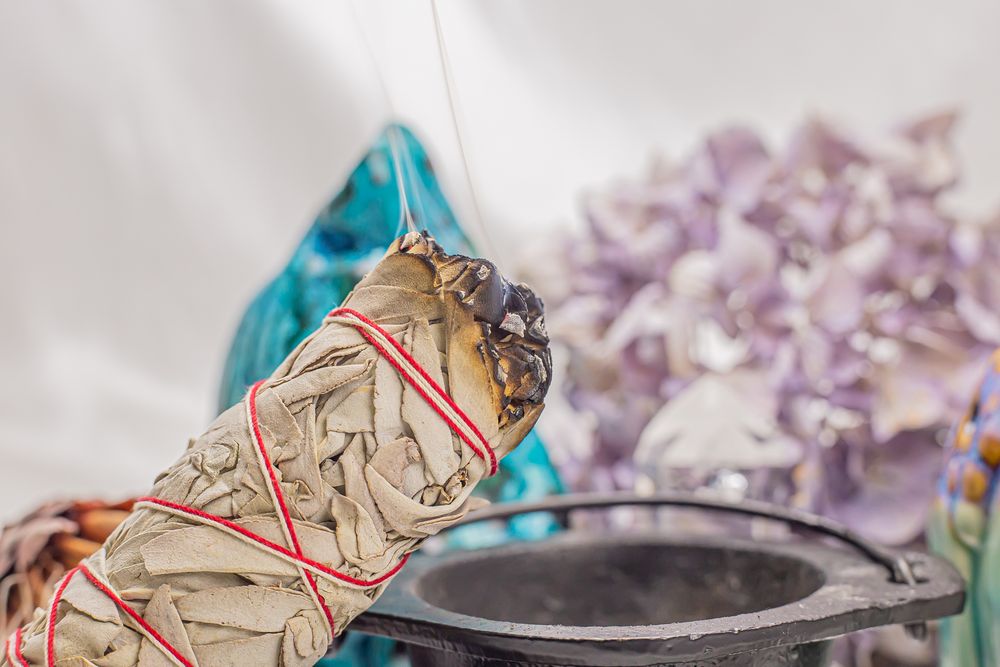 Burning sage smudge stick with smoke trails rising against a blurred purple and turquoise background.