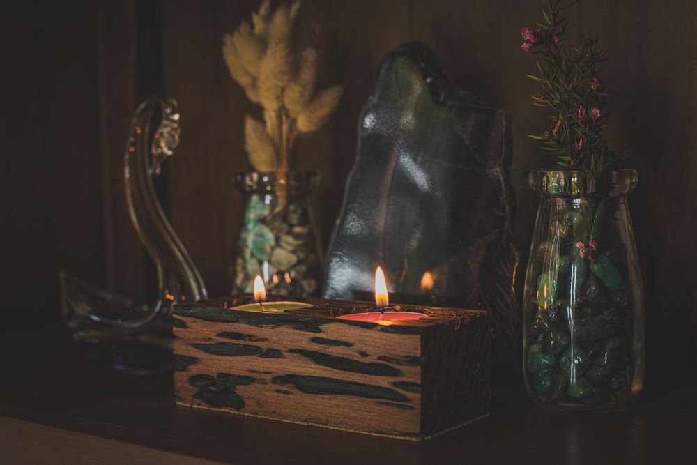 Glowing candles illuminate glass bottles and jars in a dark moody setting creating an atmospheric still life scene.