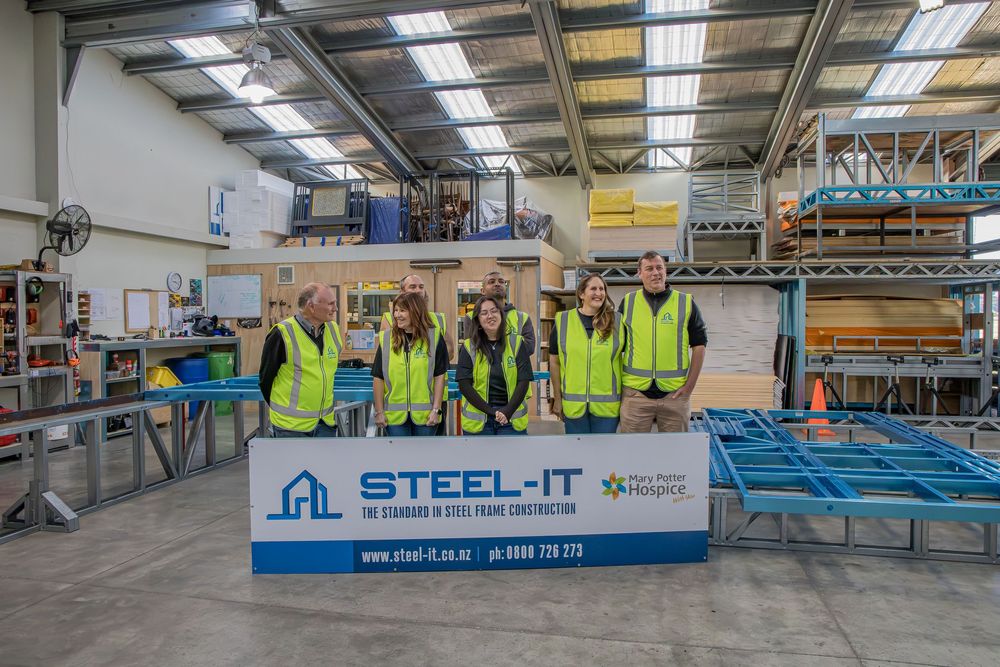Group of professionals in hi-vis vests at Steel-IT steel frame construction facility, standing by company banner in warehouse.