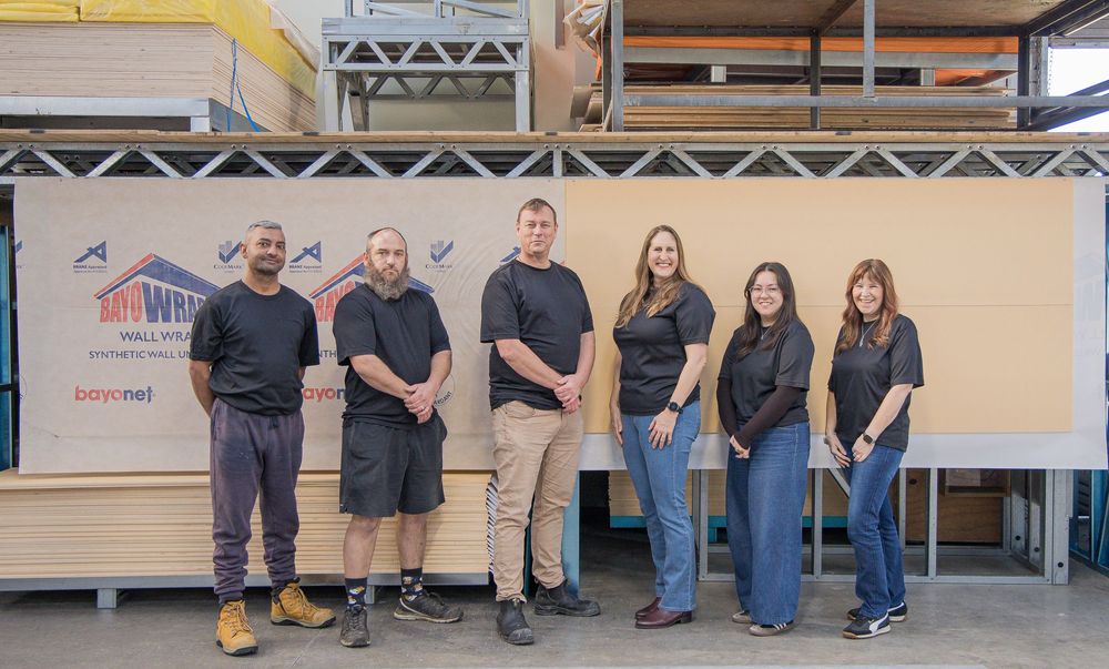 Six employees in black shirts stand together in a warehouse with building materials and company signage.