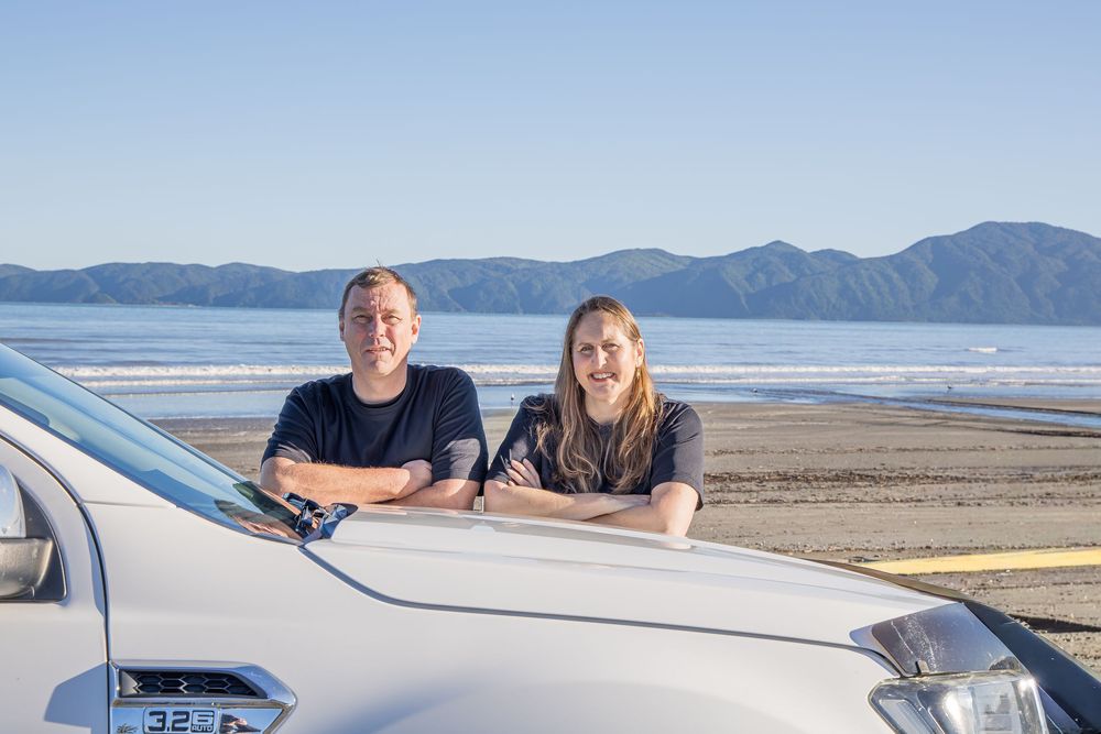 Two people leaning on a white car hood near a scenic beach with calm waters and mountains in the background.