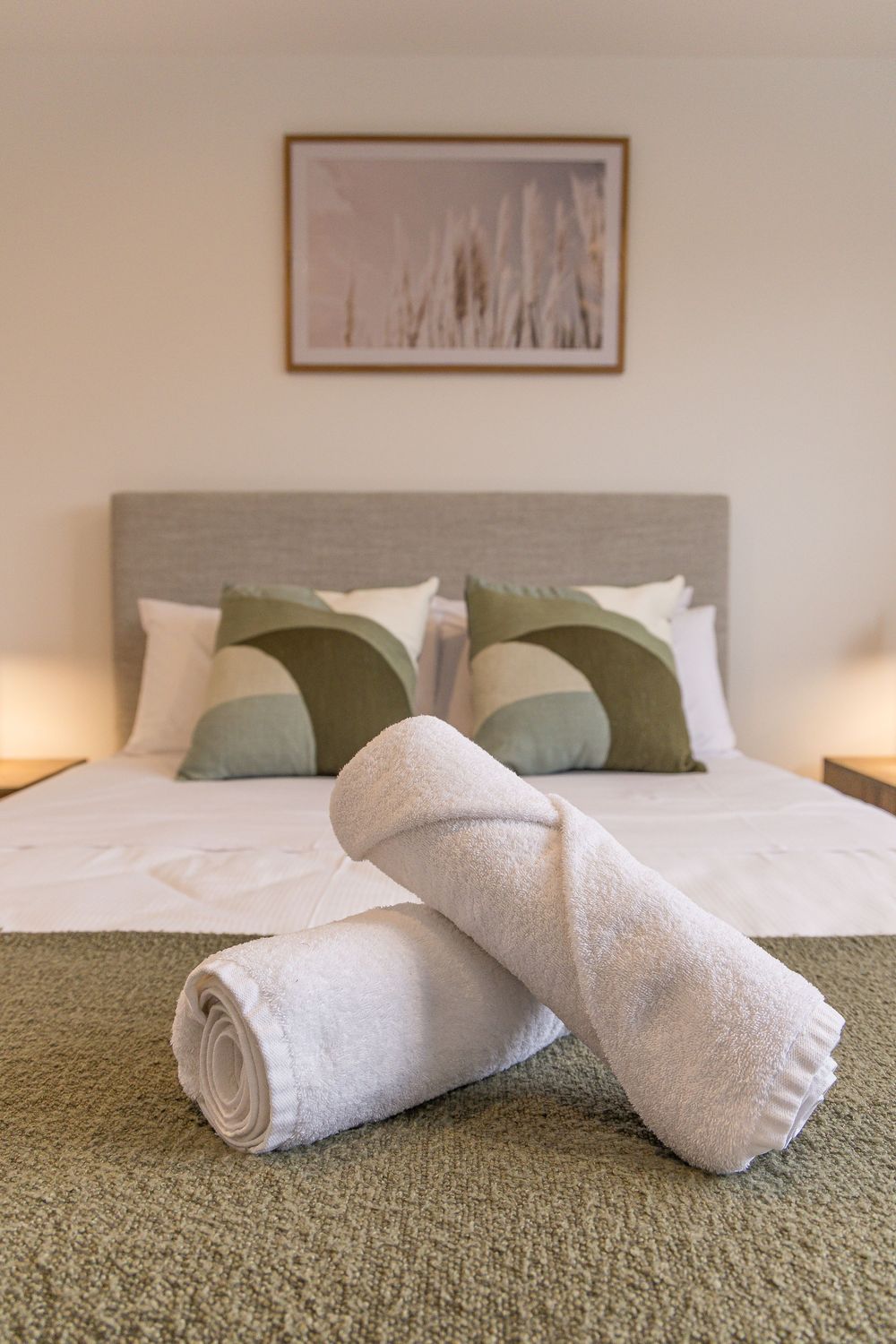 Two rolled white towels on a green carpet in front of a neatly made bed with sage green pillows.