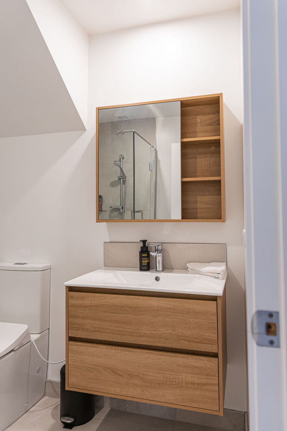 Modern bathroom with wooden vanity, mirror cabinet, white sink, soap dispenser, and glass shower visible in reflection.