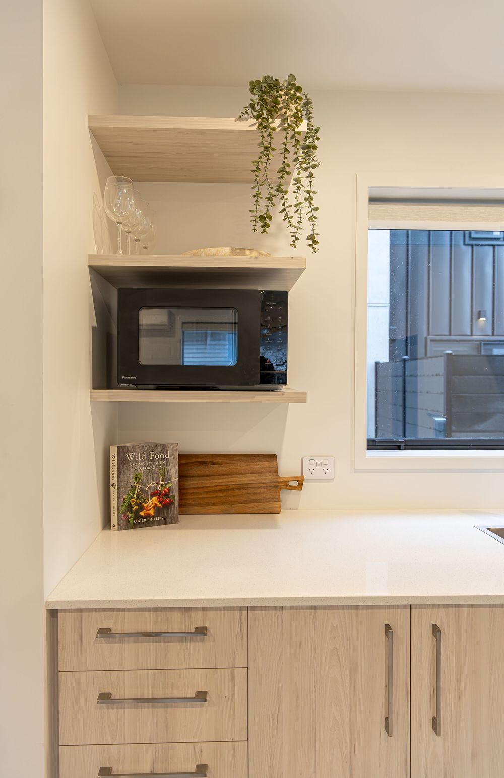 Modern kitchen corner with light wood cabinets, wall-mounted shelves holding a microwave, cookbook, and trailing green plant.