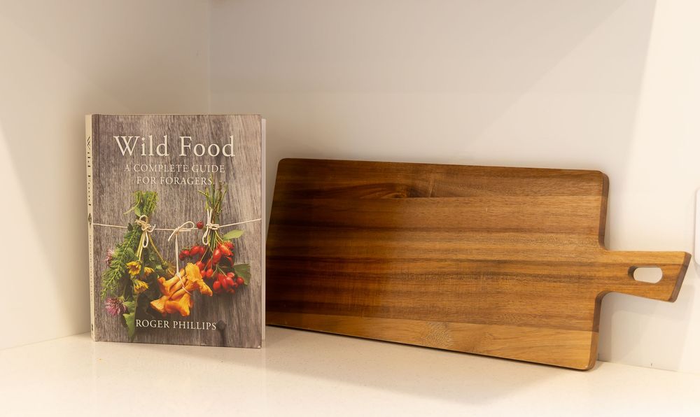 Wild Food book by Roger Phillips beside a rustic wooden cutting board with handle on a white shelf.