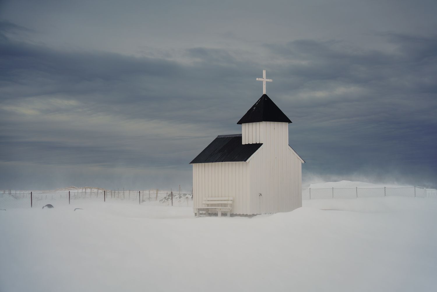 Small white church with black roof and cross stands alone in a vast snowy winter landscape under cloudy skies in Harstad, Lofoten Islands.