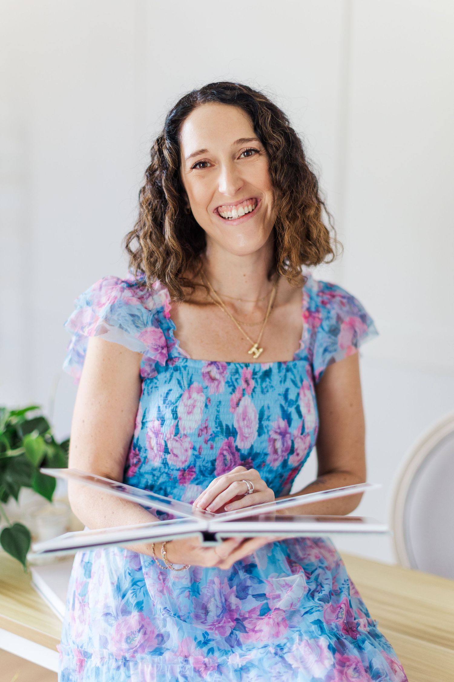 Professional photographer in casual attire sits with camera on pink chair near houseplants in bright indoor setting.