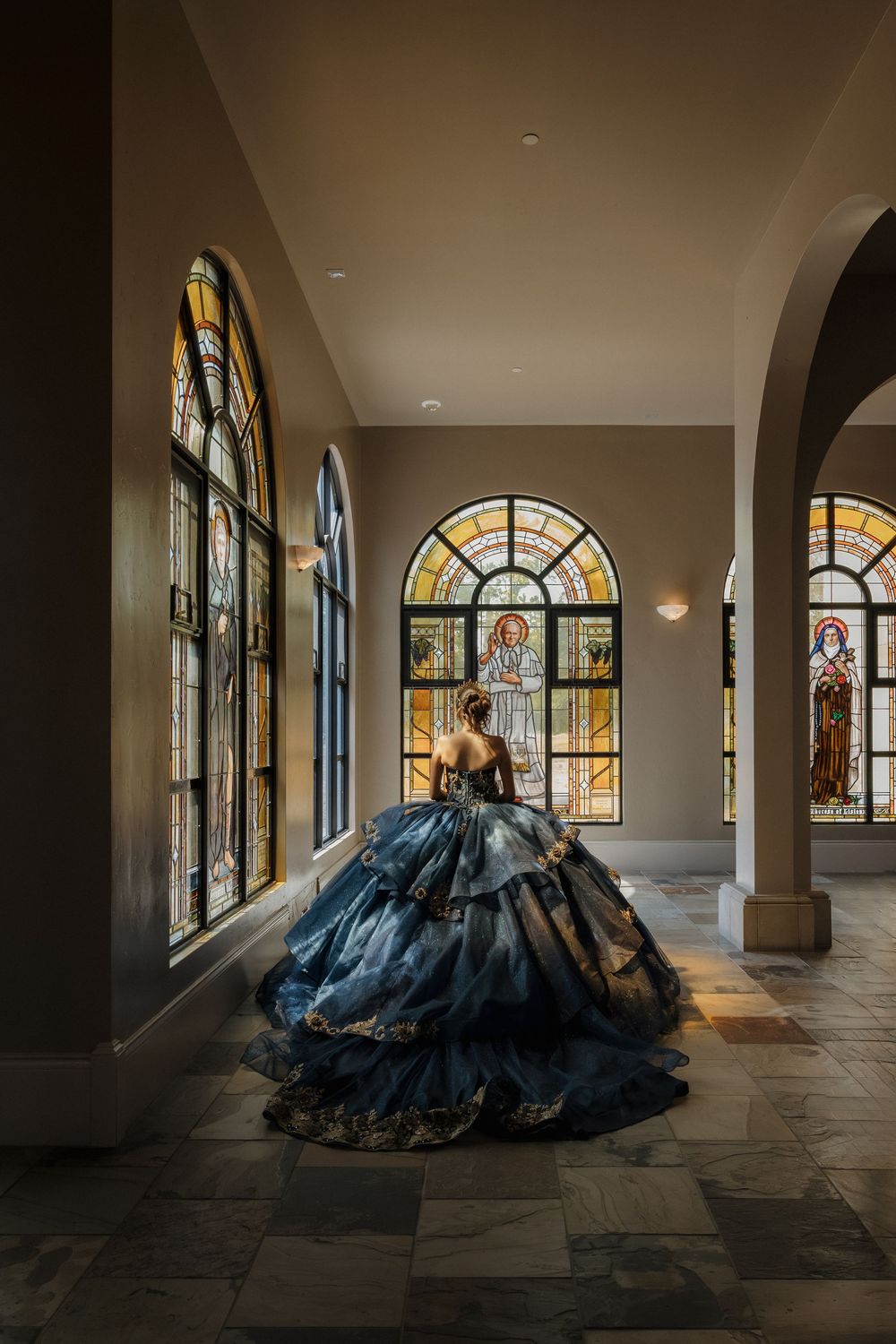 A dramatic blue ball gown sweeps across a hallway with stained glass windows in a grand architectural setting.