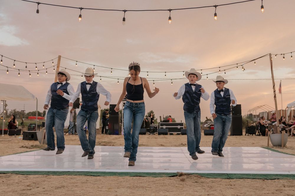 A row of people in denim and boots stand at sunset with string lights overhead creating a rustic outdoor ambiance.