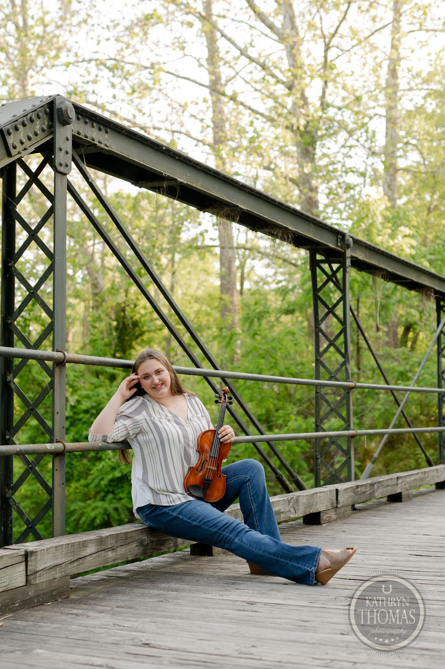 Violinist poses with instrument on wooden footbridge in natural outdoor setting.