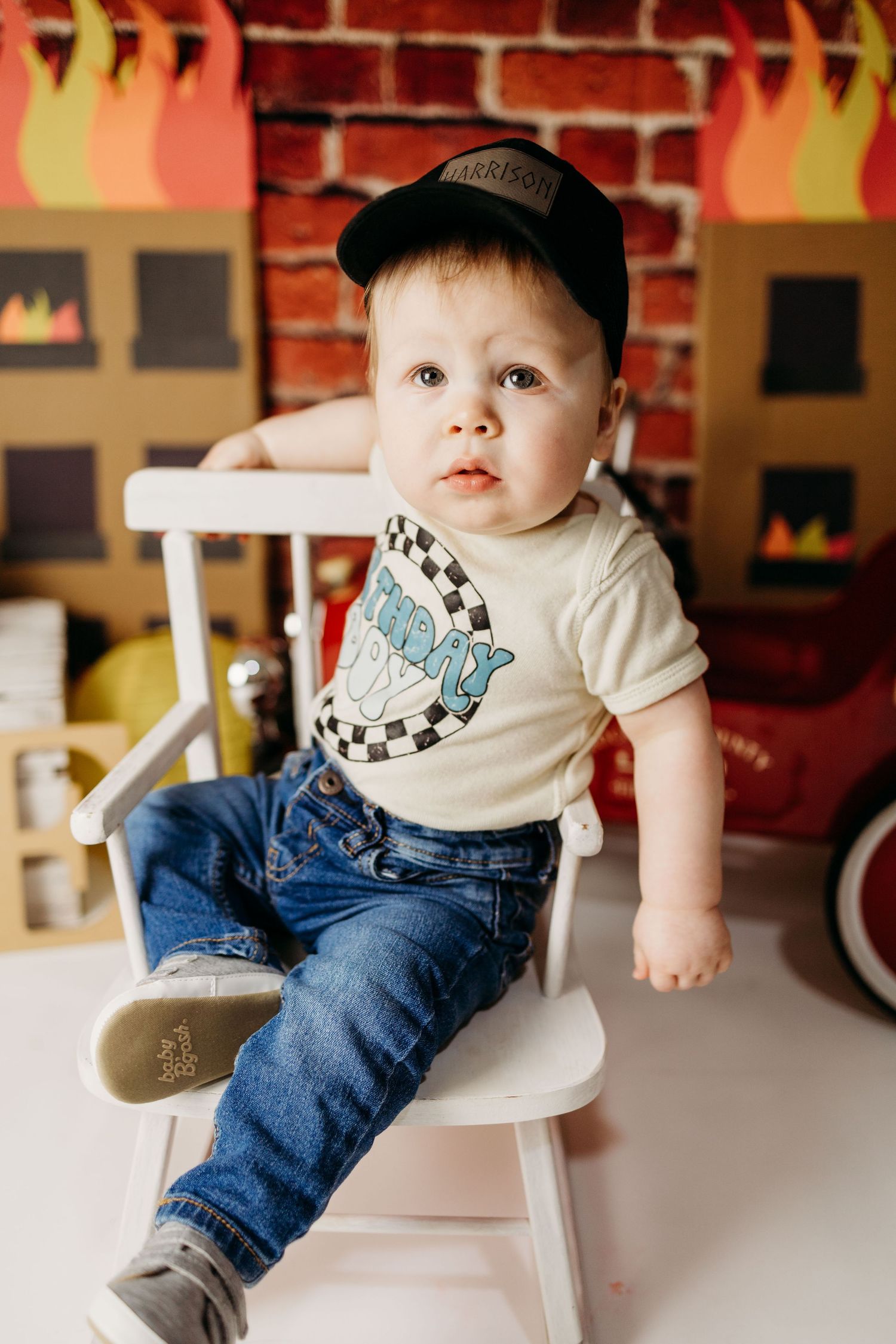 A child in a cap and graphic t-shirt sits in a white chair against a brick wall decorated with paper flames.