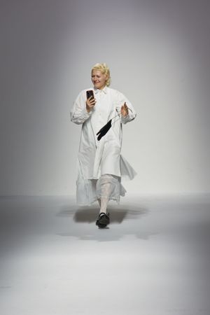 Model walking on runway wearing an all-white ensemble while looking at phone.