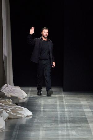 Designer waving on runway wearing all black outfit during fashion show finale.