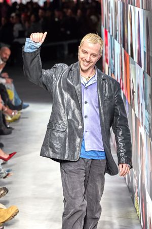 Designer in metallic jacket and jeans waves to audience during fashion show.