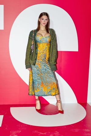Model wearing striped green cardigan over yellow-blue patterned dress against red backdrop.