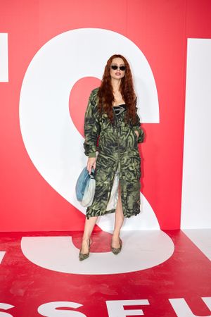 Model in camo print dress and sunglasses poses against bright red wall with white geometric design.