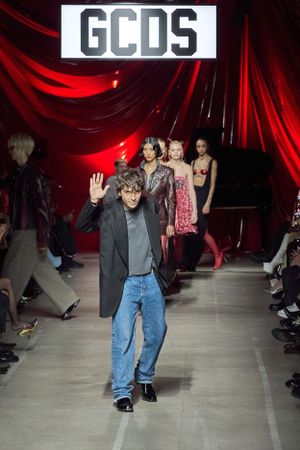 Designer waves to audience during GCDS fashion show finale on runway with red curtain backdrop.