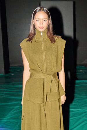Model wears olive green zip-front dress with belted waist against emerald backdrop.