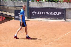 Tennis player walks on clay court with Dunlop banner visible in background.