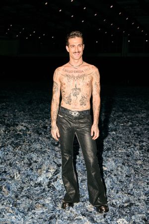 Model with tattoos poses shirtless in leather pants against dark atmospheric background.