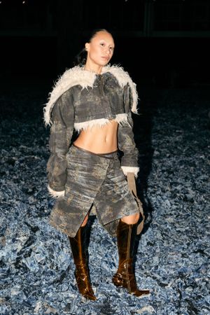 Model in distressed denim and fur-trimmed jacket poses with brown boots on textured floor.