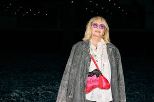 Model wearing grey coat and sunglasses poses against dark background with starry lighting.